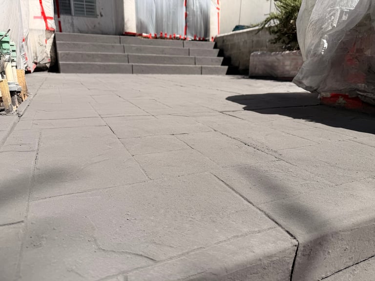 Concrete patio or plaza with gray paving stones, stairs in background, construction materials and plastic sheeting visible