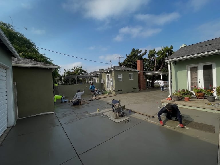 Residential neighborhood street with workers finishing concrete driveway installation in front of small houses under blue sky